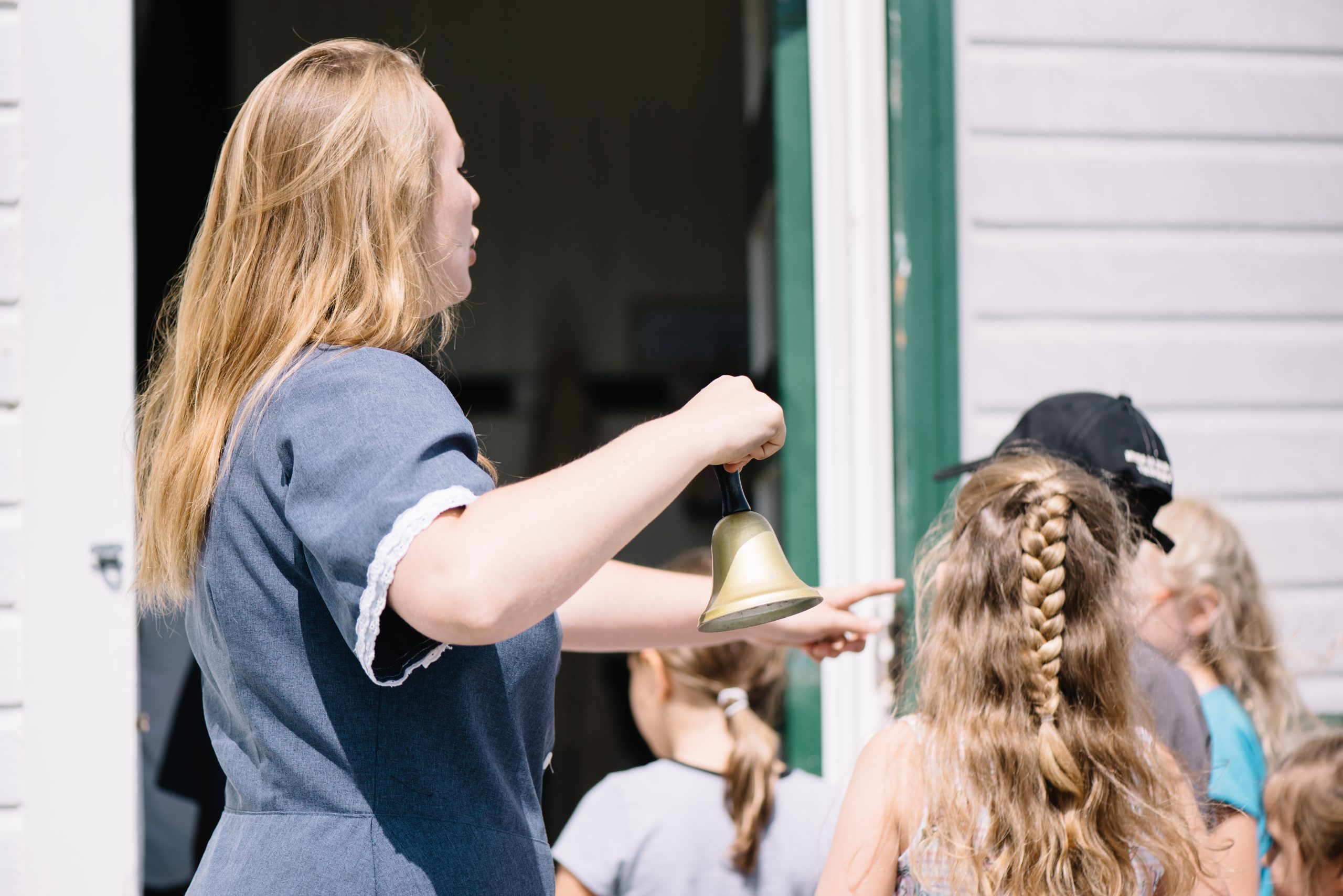 Une femme au long cheveux blond tient une cloche. Des enfants entrent dans l'école du rang II. Visiter ce musée-école est une belle activité à faire en famille en Abitibi-Témiscamingue. 